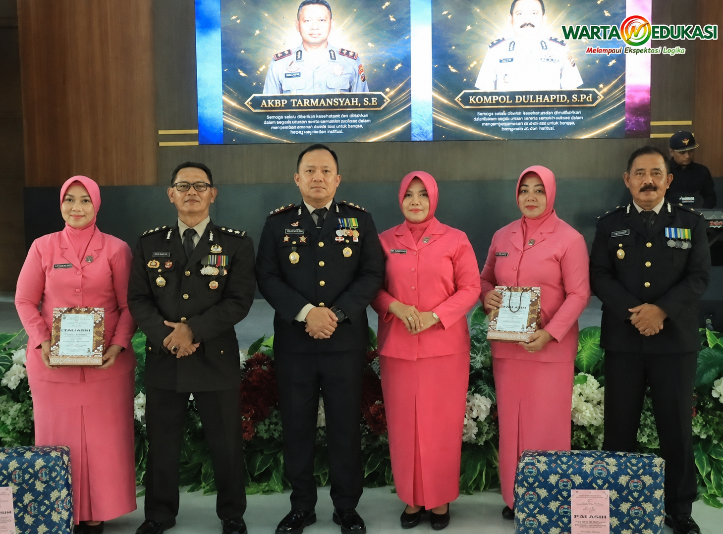 "Enam peserta acara, terdiri dari personel kepolisian dan anggota Bhayangkari, berpose bersama dalam acara seremonial resmi di dalam ruangan dengan logo Warta Edukasi di pojok kanan atas."