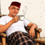 Seorang pria mengenakan kaos oblong putih dan peci merah maroon yang sedikit miring, duduk santai di kursi bambu sambil memegang secangkir kopi panas.